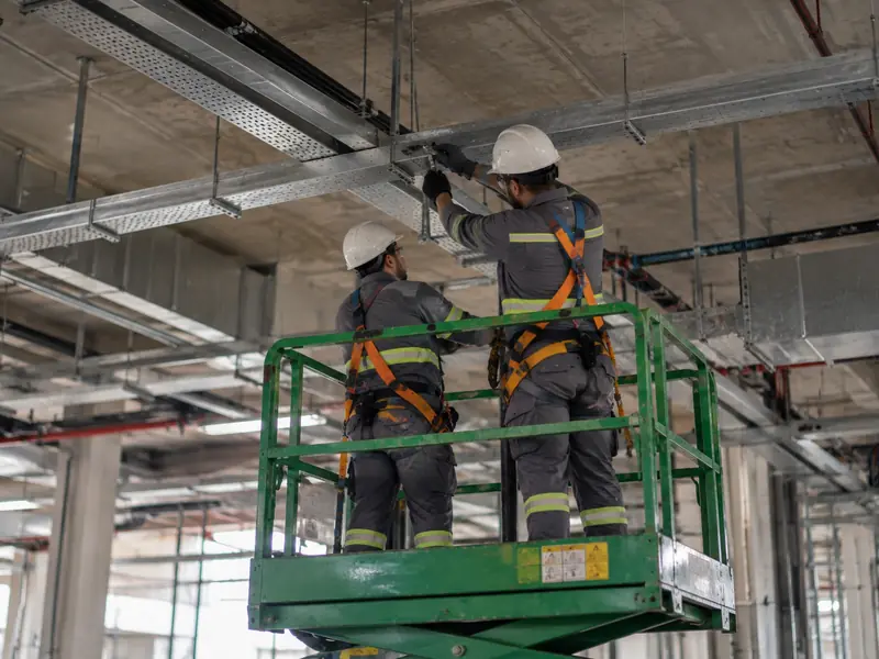 Dois técnicos com cinto de segurança em plataforma elevatória instalando eletrocalha