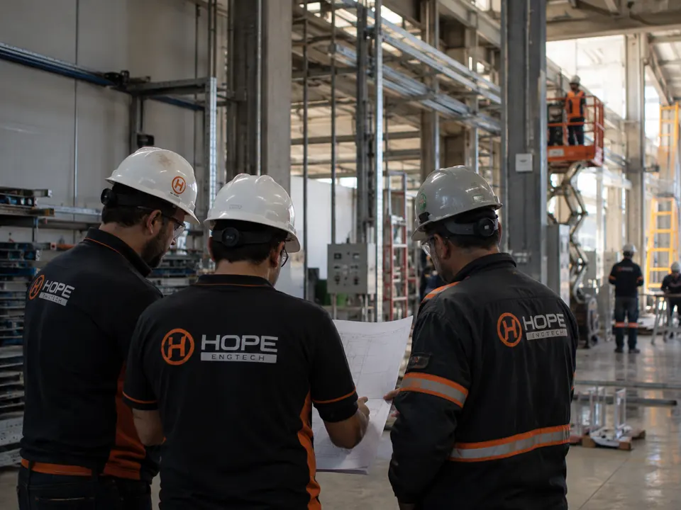 Três profissionais com uniforme HOPE ENGTECH e capacete analisando uma planta em um galpão industrial